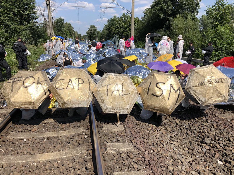 [The golden finger sits on tracks and holds umbrellas in the air that say "FCK CAPITALISM".