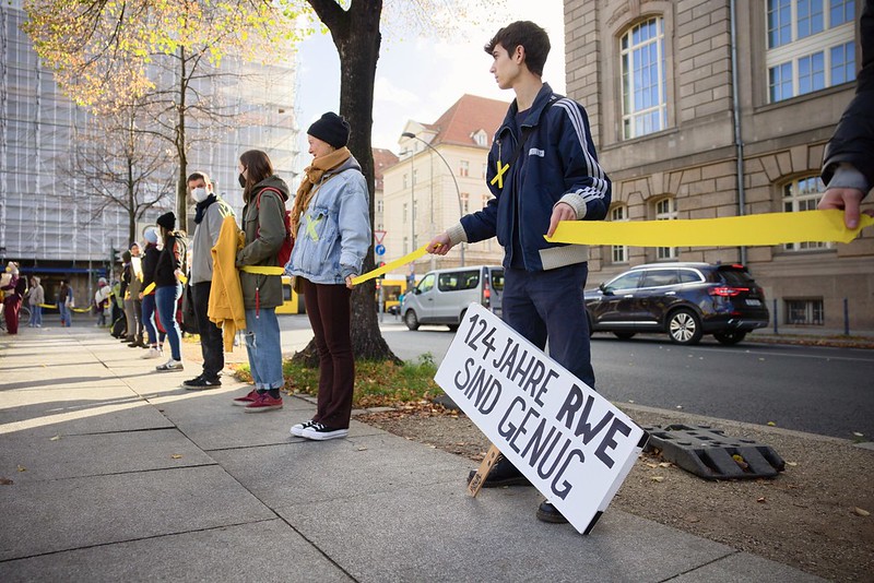 Activists stretch a yellow ribbon around Lützerath, a sign reads "1124 years of RWE are enough"