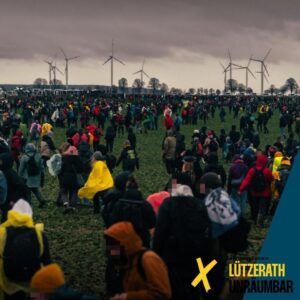 Many demonstrators with rain gear and backpacks on a field. In the background, Lützerath, police tubs and wind turbines. Grey, cloudy wind. Logo Lützerath Unräumbar